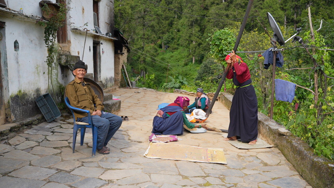 Village courtyard in Uttarakhand where women clean and pound grain while a man sits nearby