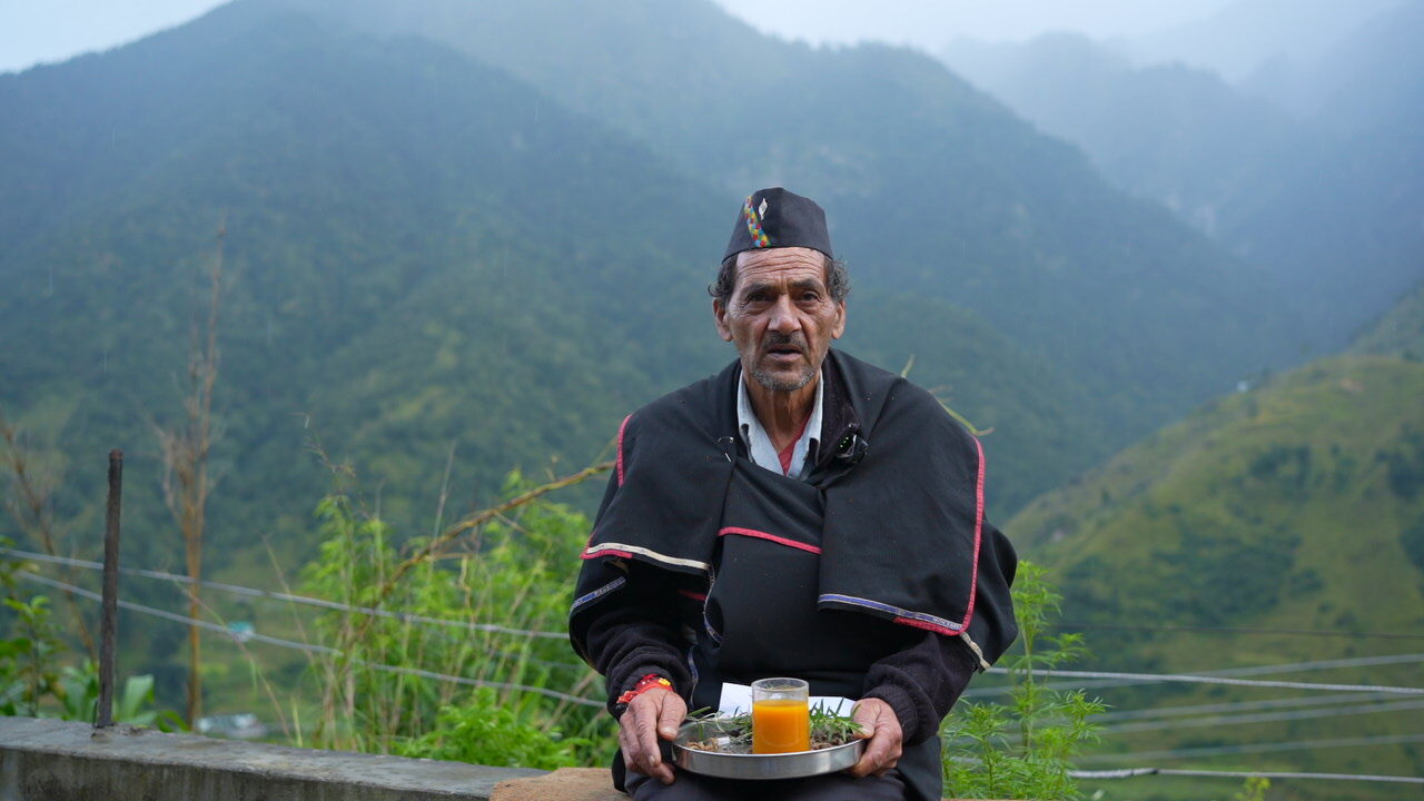 A village elder in a traditional Kumaoni cap and black wool shawl, seated with a plate of herbs and a glass of orange juice, misty green mountains behind