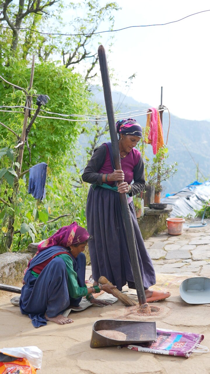 Two women pounding grain with a long wooden pestle in a village courtyard in Uttarakhand