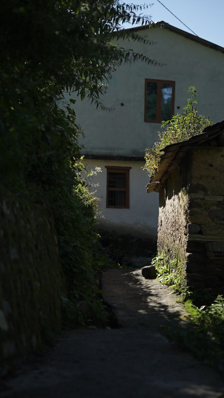 A narrow dirt lane between a stone wall and a slate-roofed stone outbuilding, leading to a whitewashed village house