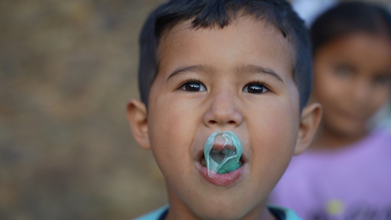 A young boy blowing a green bubble-gum bubble while another child stands blurred behind him