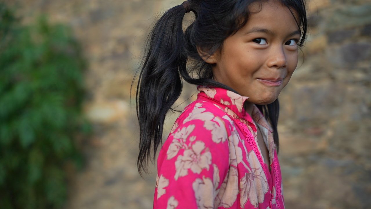 A girl in a pink floral top with a high ponytail looking back over her shoulder at the camera, in front of a stone wall
