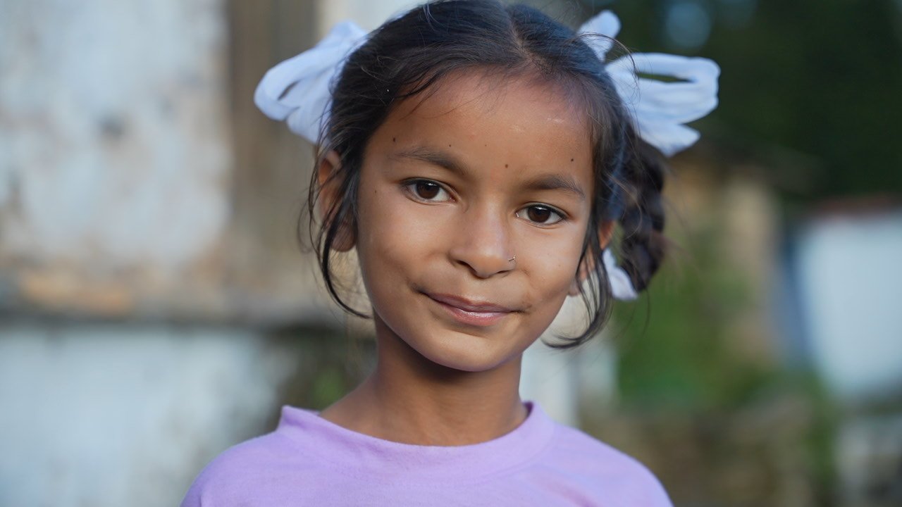 A girl with white ribbons in her hair, wearing a lilac top, looking at the camera